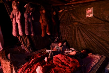 Two boys lie on a mattress inside a room with walls made of tarpaulin. Four teddy bears hang from the ceiling