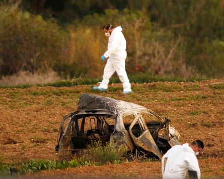 Two forensics officers in white hazmat suits search a field around the burnt-out shell of car
