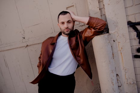 Penn Badgley in brown leather jacket, white T-shirt and black trousers leaning against an industrial pillar