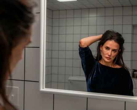 A woman with long hair looks at herself raising her arm to her head in the mirror in a tiled bathroom
