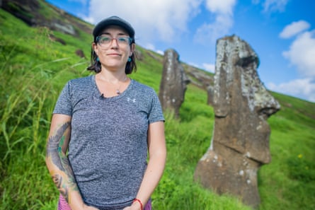 A young Hispanic-looking woman stands on a hillside with colossal statues behind her