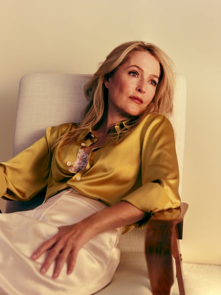 Gillian Anderson lying back in a cream armchair in front of a cream wall, wearing a white silky skirt and an unbuttoned ochre silk shirt with a white lacy bra underneath