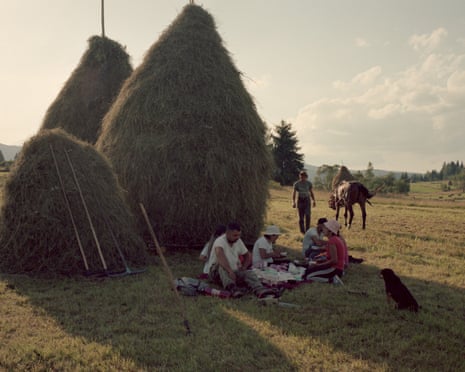 A group of people have a picnic in the late afternoon sun in the shade of a large haystack. A horse is in the background