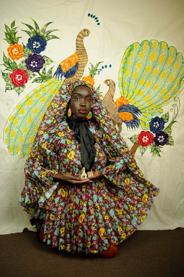 A woman in an elaborate floral headscarf and gown holds a small model house, before a painted bird mural