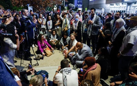 At night, outside a well-lit building, a circle of dozens of ceasefire delegates and press, those in the middle sitting and those in the back standing.