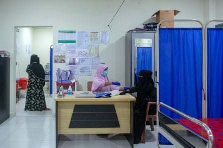 A woman in a pink chador sits behind a desk measuring another woman’s blood pressure in a new and modern-looking hospital room.