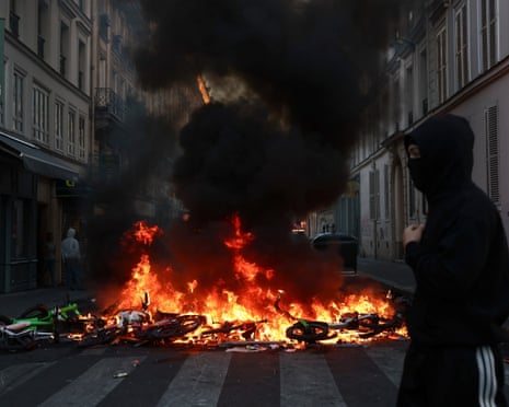 A masked person stands near burning bicycles during civil unrest in Paris
