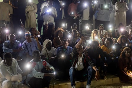 A group of young Africans sit in rows at night holding torches and candles