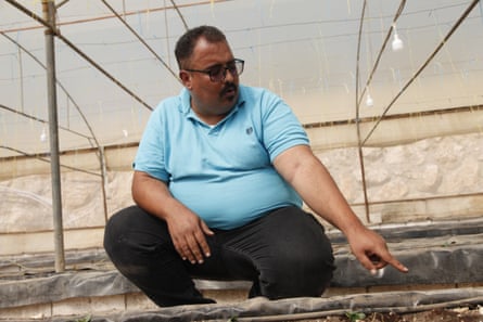 An overweight middle-aged man sits in a greenhouse pointing at rows of seedlings