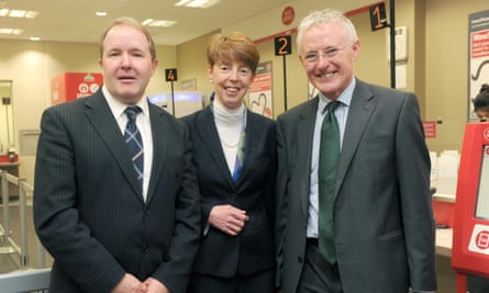 Paula Vennells, with short hair and wearing a turtleneck shirt and jacket with a strand of pearls, stands smiling next to George Thomson and Norman Lamb, both in suits and ties, in a post office