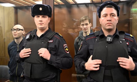 Artyom Kamardin and Yegor Shtovba in a glass cage with two security officers or police in front of them