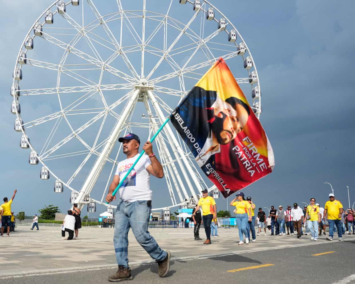 A man holding a flag on a pole depicting Espriella walks with others past a ferris wheel