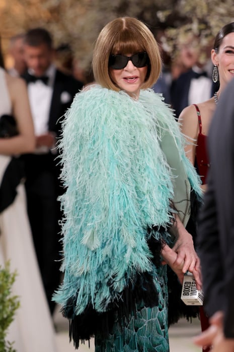 woman with black sunglasses and teal feathered cape poses