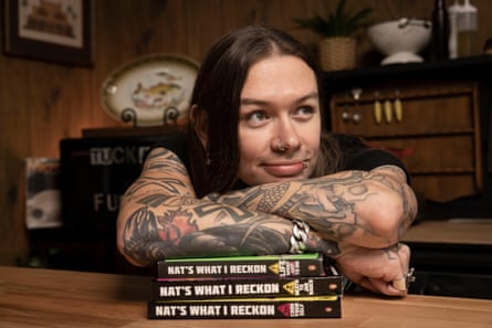 A man with. long hair and arm tattoos resting his arm on a stack of books.