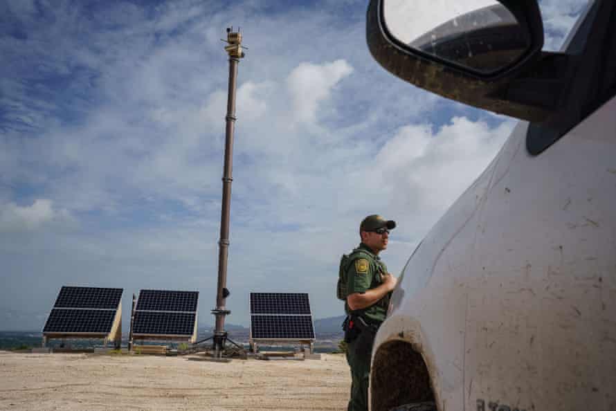 A border patrol agent stands near to three solar panels being used to power a portable surveillance camera installed on a tall pole.