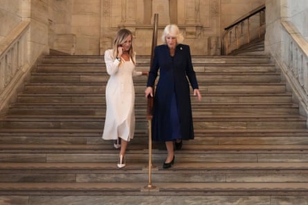 A woman in a white dress, and a woman in a dark blue dress, descend a stone staircase together with the banister between them.