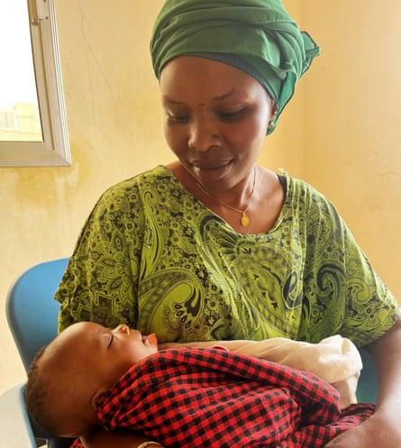 A young Senegalese woman in traditional dress, holding and looking down with a smile at her sleeping baby.