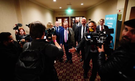 Nigel Farage is surrounded by members of the media and security in a hotel corridor