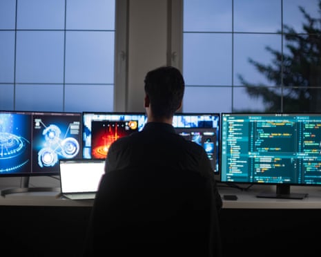 An IT engineer sits facing monitors displaying code and graphics in a dim room