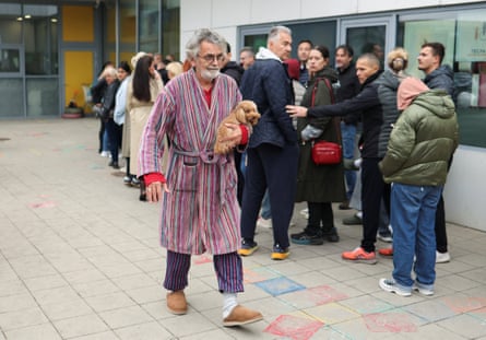 A man in a striped robe carries his dog past a queue outside a polling station