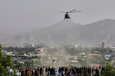 A military helicopter drops flowers over Kabul as a group of men stand on a hillside in what looks like a park looking over the city; there are mountains in the background