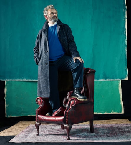 Actor Michael Sheen standing on a chair in front of a green wall hanging