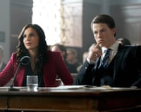 A woman and a man in suits on the front desk in a courtroom, looking very very serious, the young man stroking his chin thoughtfully