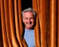 Charlie Nicholas smiles while peering out through a gap in orange velvet curtains