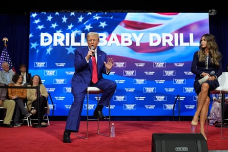 An elderly man with dyed blond hair talks on stage next to a younger woman, with the words ‘Drill Baby Drill’ on the screen behind them