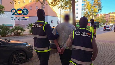 Two men in hi-vis uniforms lead a third man in handcuffs away from the camera
