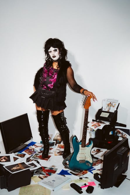 a person in white face paint poses with a guitar surrounded by monitors and photos on the floor