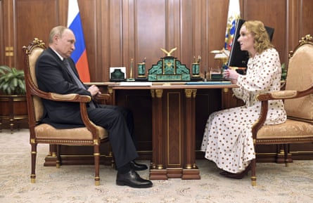 A man in a suit sits opposite a woman in a dress in a wood-panelled office with a flag behind them