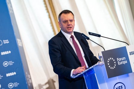 he stands at a podium in front of microphones. He is in his mid-40s with short, light brown hair and wears a dark suit, white shirt and red tie.