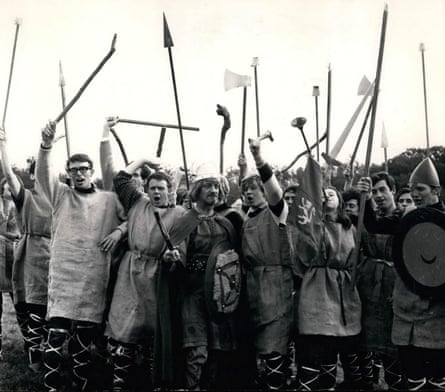 A black and white image showing a crowd of men in a field dressed in medieval combat wear raising spears and axes