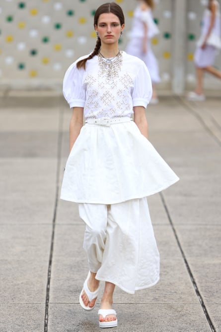 A model wearing dressed in all-white including a broderie anglaise short-sleeved top, belt and skirt, over wide-leg, ankle-length trousers