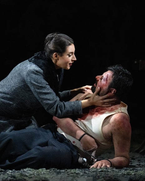 A woman in a coat with bare feet leaning over a man who is covered in blood