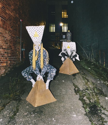 Two people dresked in polka-dot costumes wearing polka-dot headwear sitting on triangular shapes in a dark alley