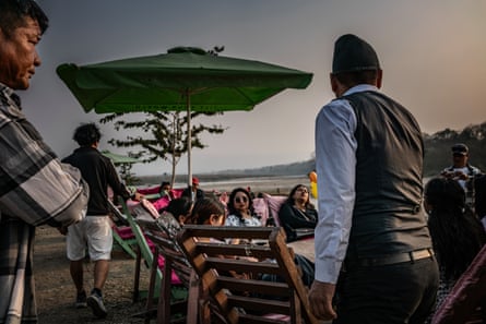 People sit in chairs with an umbrella next to them. Other people stand around them. A body of water can be seen behind them.