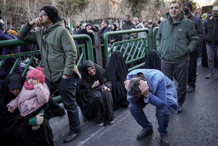 A grief-stricken man crouches with his hands over his ears in the street. Behind are other mourners including crying women in black hijabs and full-length abayas
