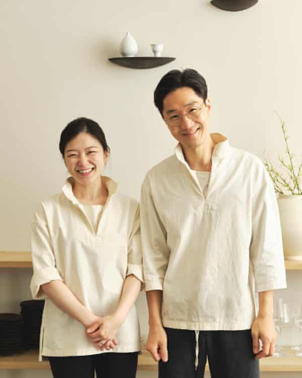 Bomee Ki, left, and Woongchul Park in matching whites standing side by side and smiling in front of a cream-coloured wall