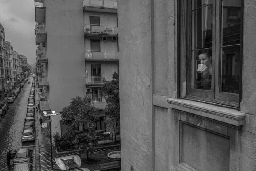 Marta at the window while drinking a herbal tea. In the background, some people queue in the street waiting to enter the supermarket below the apartment.