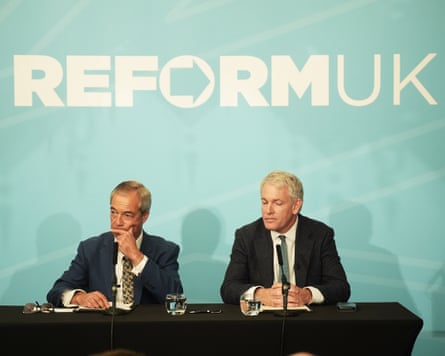 With Nigel Farage during a Reform press conference in Westminster in September this year. They are sat at a dark desk, wearing dark suits, in front of a blue backdrop featuring the words ‘Reform UK’