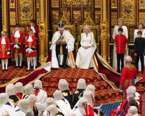 King Charles III in ceremonial robes sits on a throne before an ornate gilded backdrop, addressing an assembly below.