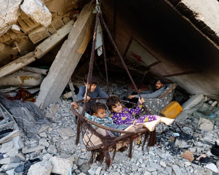 Three children play with a makeshift swing hanging from the rubble of a building. A man sits behind them among the rubble in a chair.