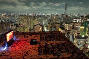 People sit in deckchairs watching a film on a rooftop with the lights of the city receding into the distance behind them