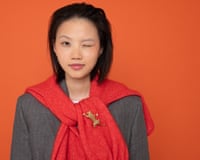 A young woman with a dark bob waring a grey jacket with a red cardigan tied around her neck like a scarf, with a leopard brooch pinned to it