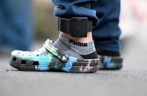 Closeup of black ankle monitor on feet of a man who is wearing multicoloured Crocs footwear and grey Puma socks