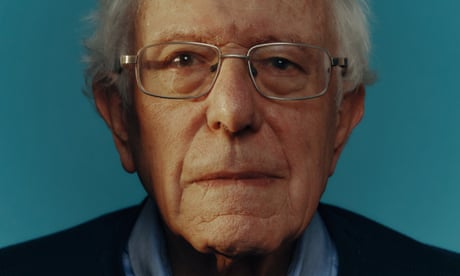Bernie Sanders wearing glasses, a blue shirt and navy jumper, against a blue background