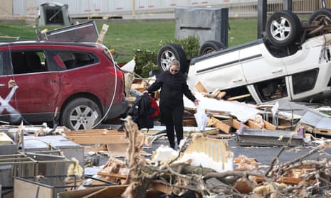 A woman holds items amid debris and an overturned car