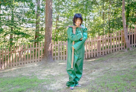 Mia, a girl on the autism spectrum, photographed in a green rubber suit, holding a pickaxe, with trees behind her, 2016